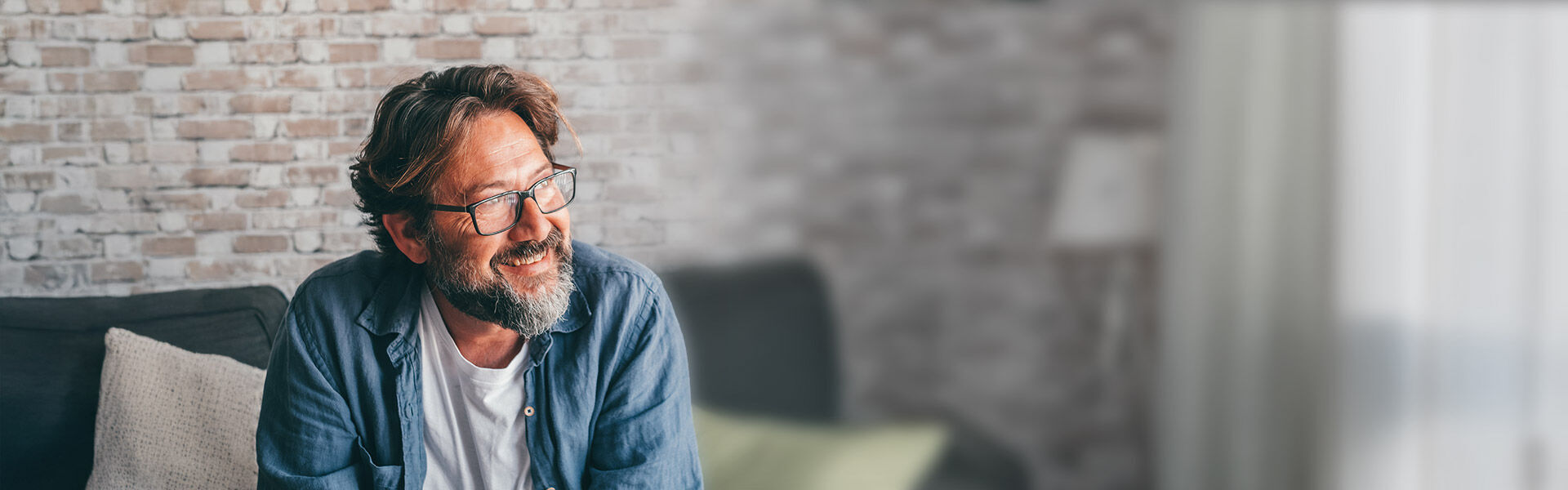 Mann sitzt im Wohnzimmer auf einem Sofa, schaut nachdenklich.